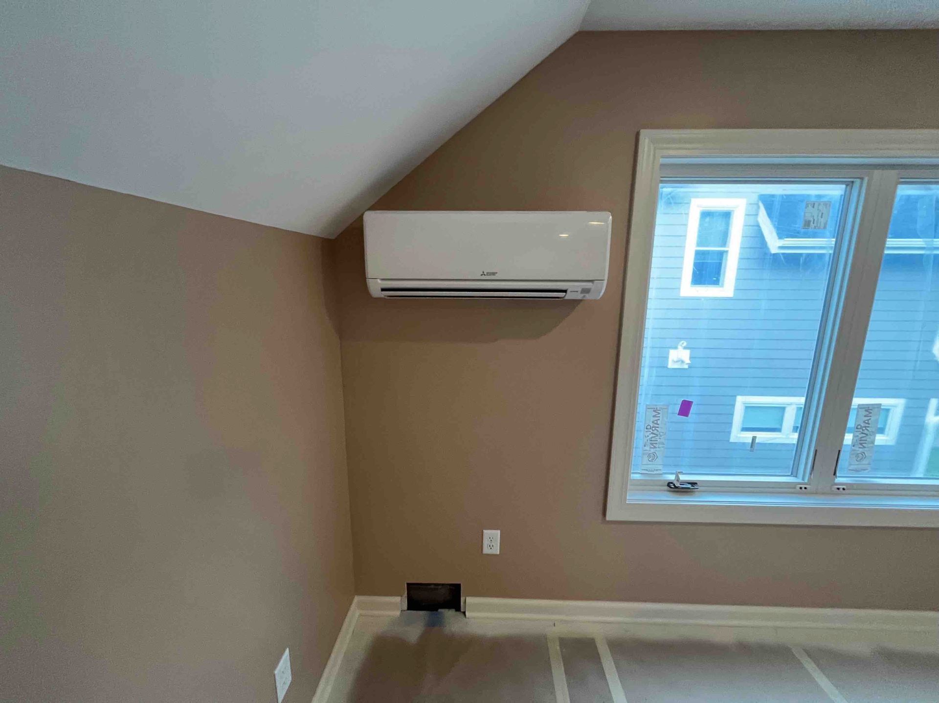 Air conditioning unit on a beige wall in a room with a window, baseboard, and angled ceiling.