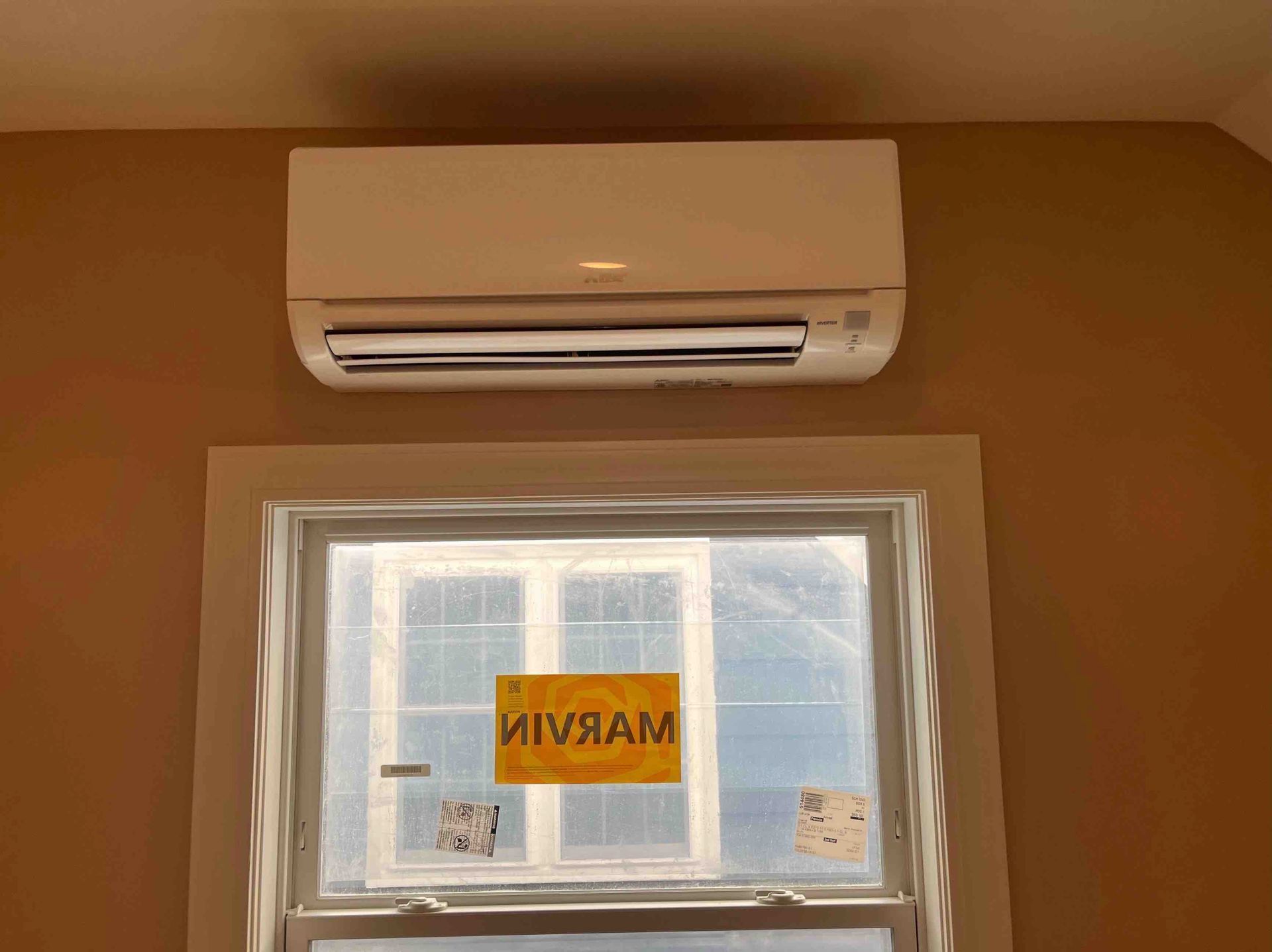 White ductless AC unit above a window, set in a tan-painted room.