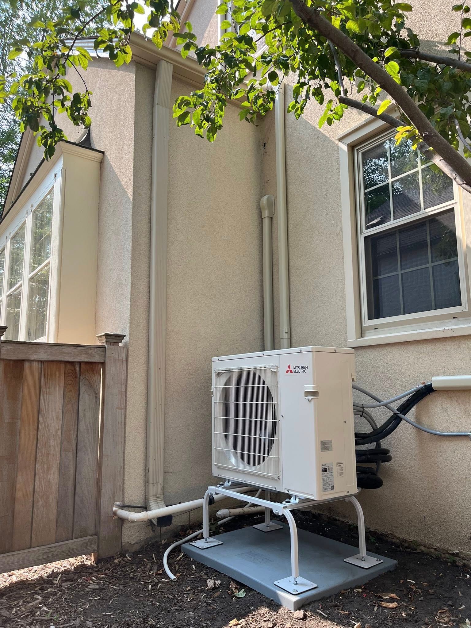 An outdoor heat pump unit mounted on a platform against a stucco building, near a window.