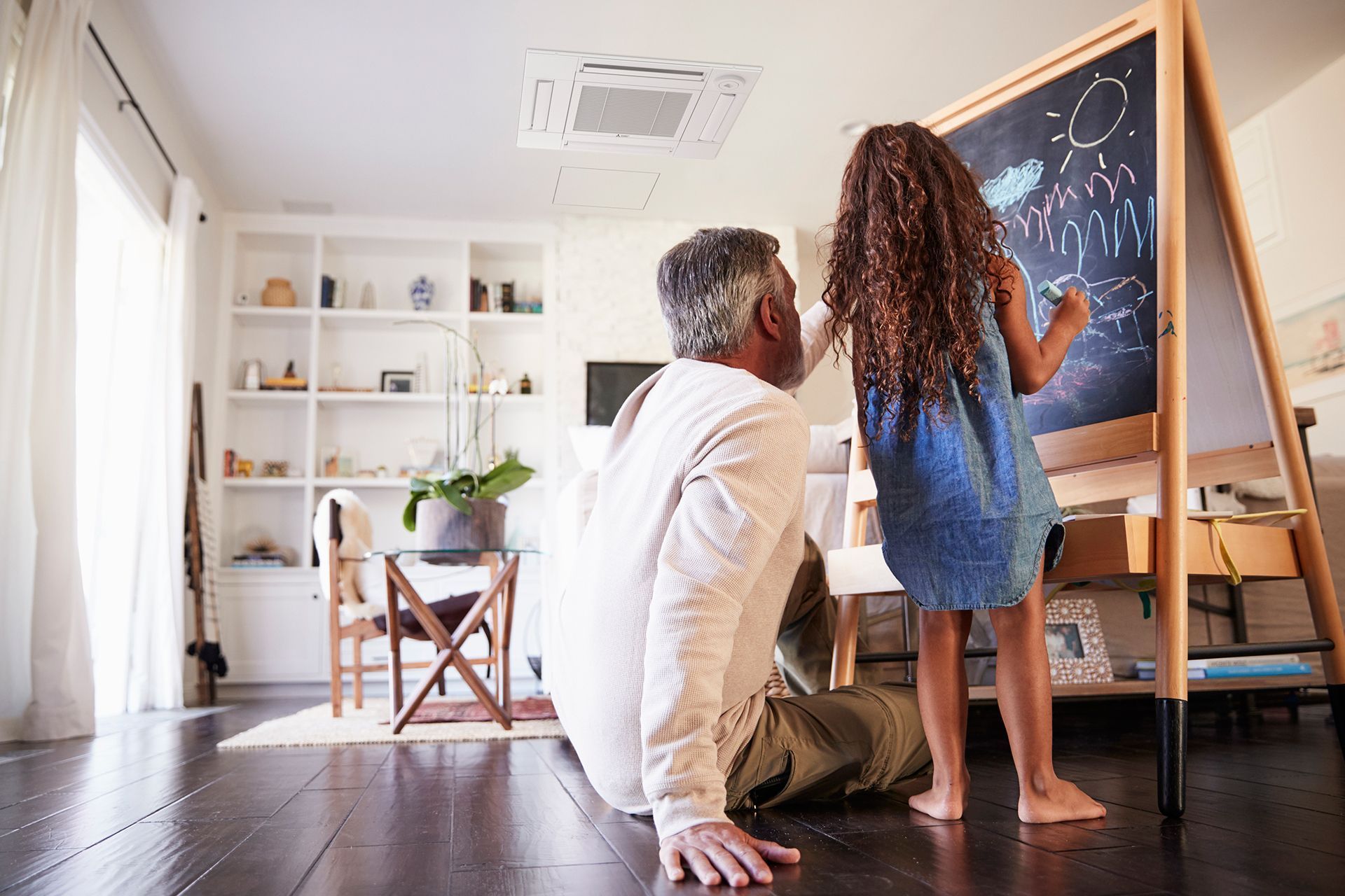 A person watches a child draw on a chalkboard easel indoors.