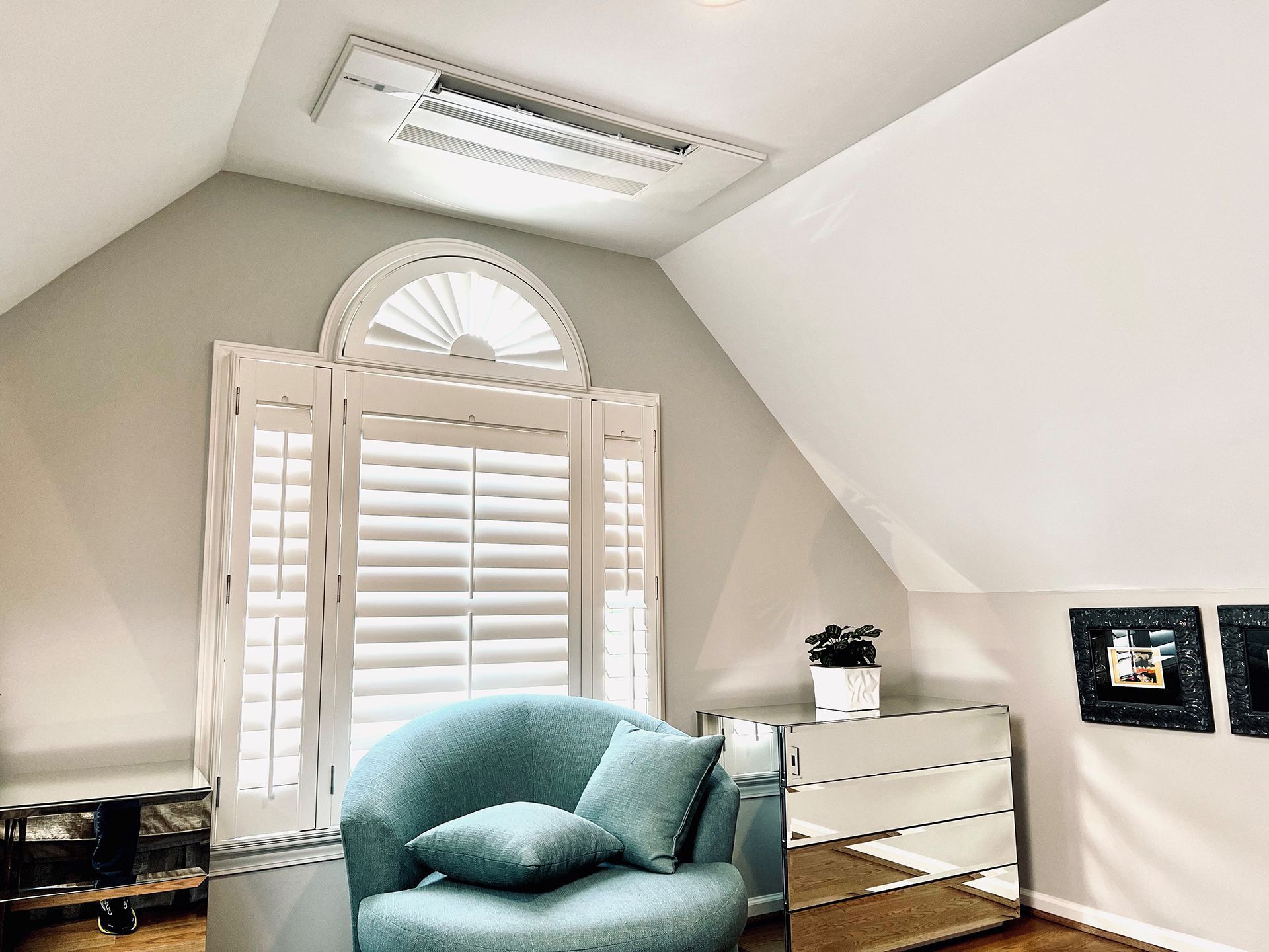 Bright room with arched window and shutters, blue chair, mirrored dresser, and ceiling lighting.