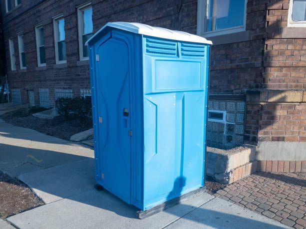 A blue portable toilet is parked on the sidewalk in front of a brick building.