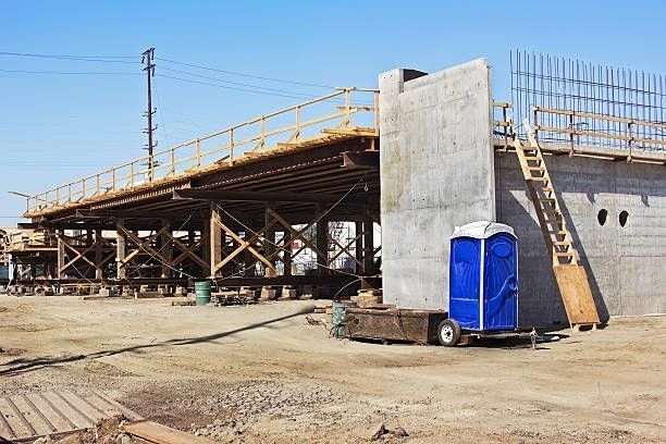 A blue portable toilet is parked in front of a bridge under construction.