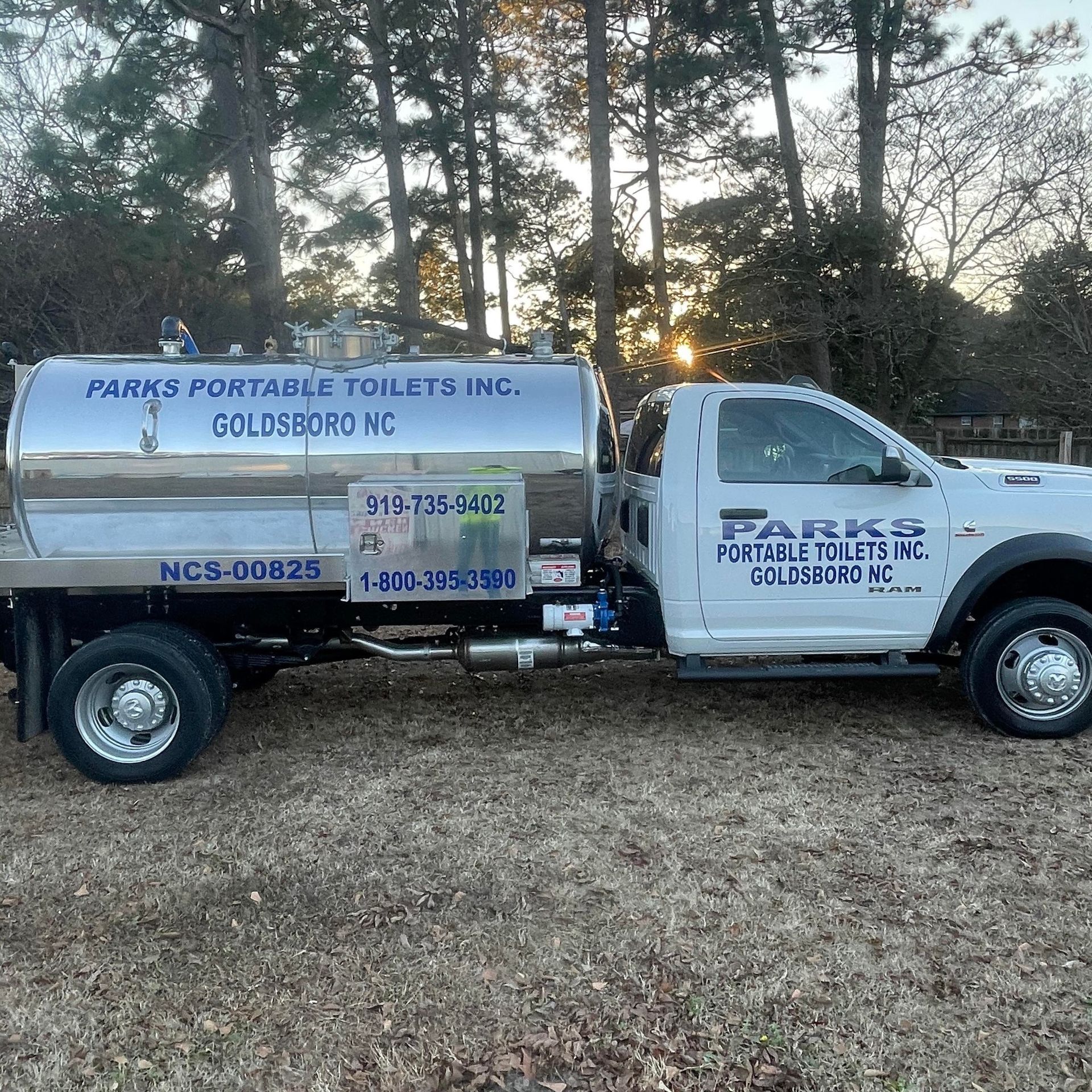 A white septic truck is parked in a field with trees in the background