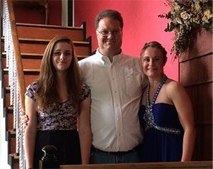 Man with two young women smiling in front of a staircase, against a red wall.