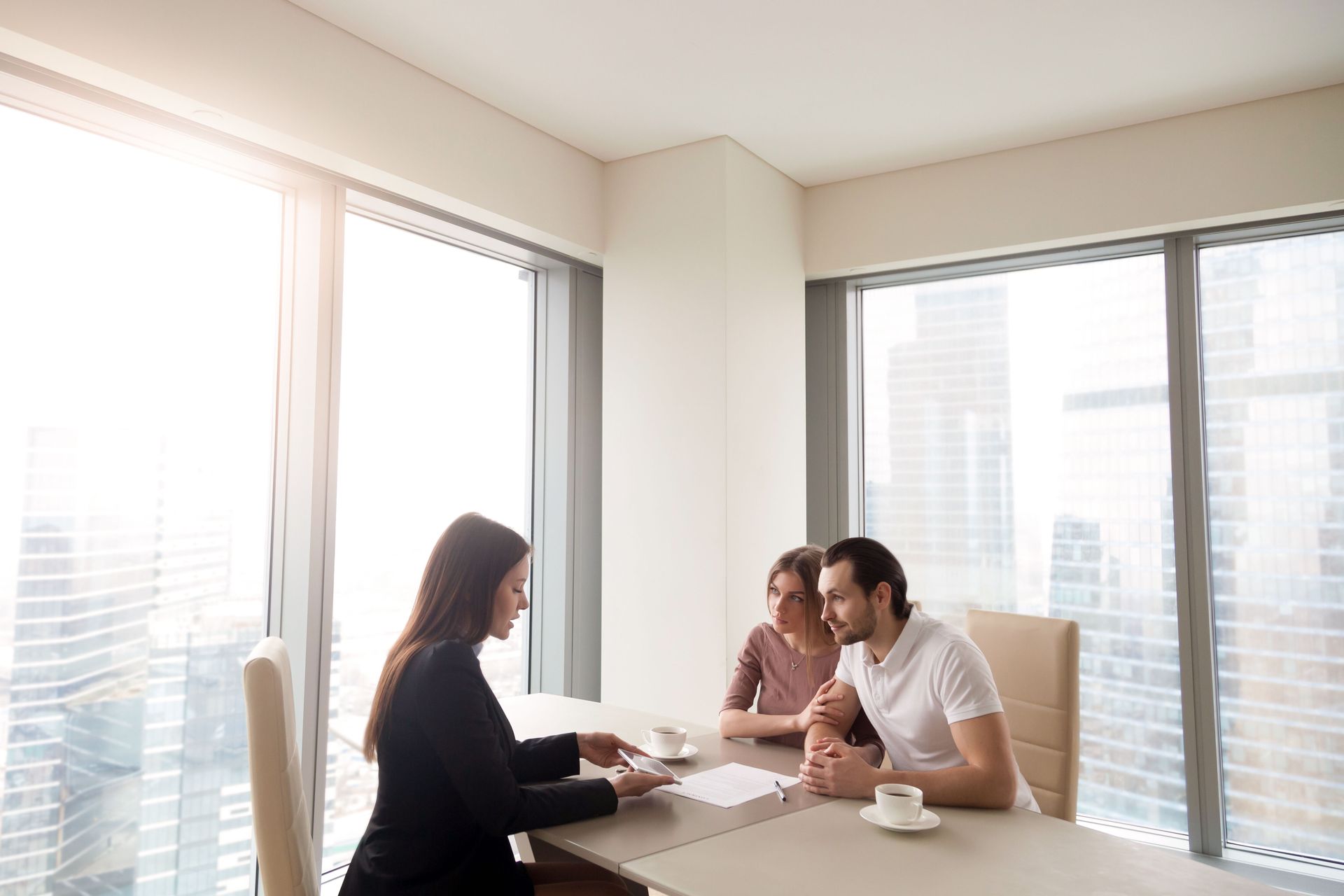 A woman in a suit reviews papers with a couple at a table near a large window.