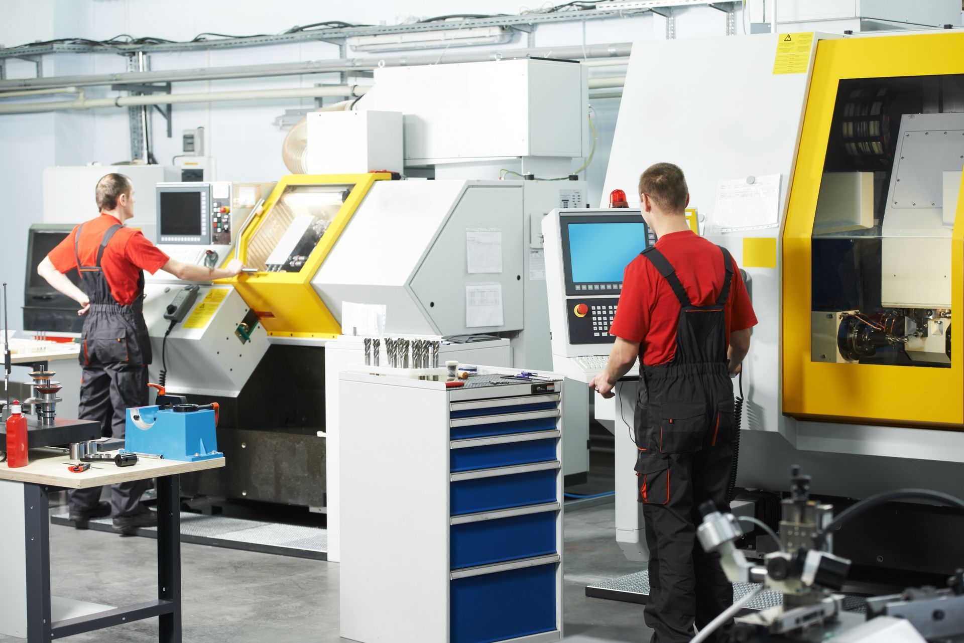 Two workers in red shirts and overalls operate machinery in a workshop, yellow and white machines.