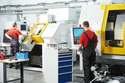 Two workers in red shirts and overalls operate machinery in a workshop, yellow and white machines.