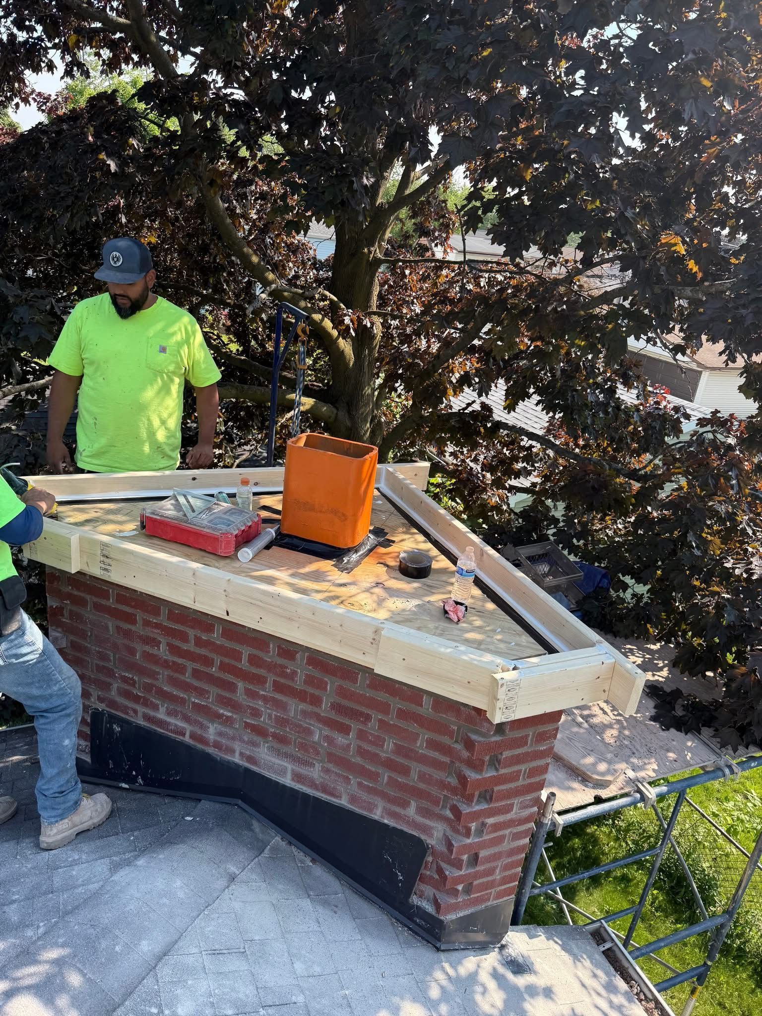 Two men are working on a brick chimney on a roof.