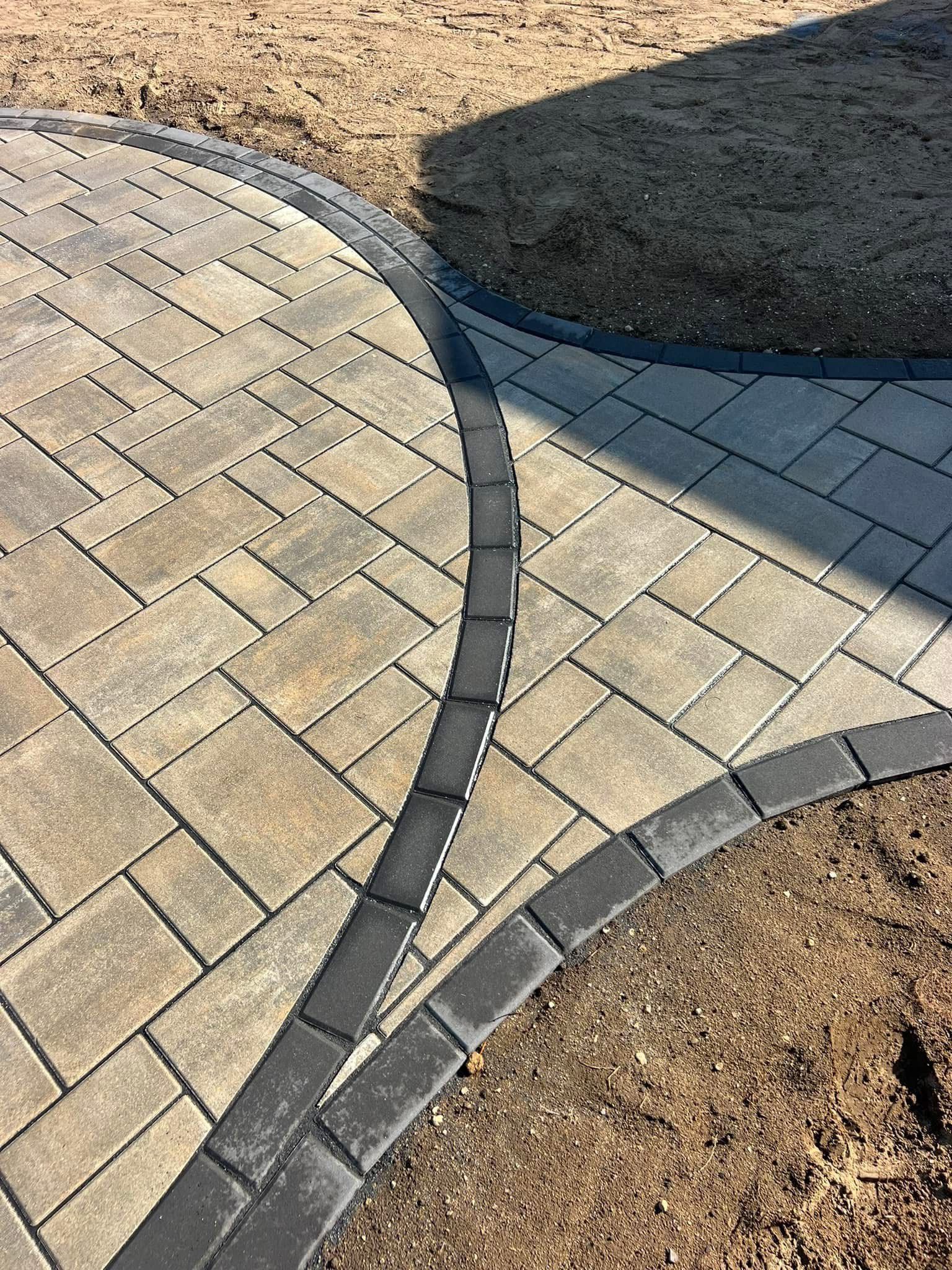 A brick walkway with a black border is surrounded by dirt.