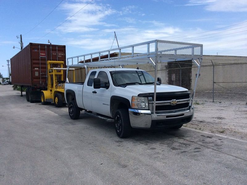 White pickup truck towing a yellow trailer with a red shipping container on a street beside an industrial building