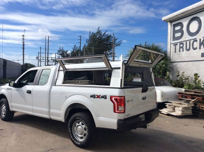 White pickup truck with camper shell parked beside a building with stacked lumber and a sign reading “BOX TRUCK”