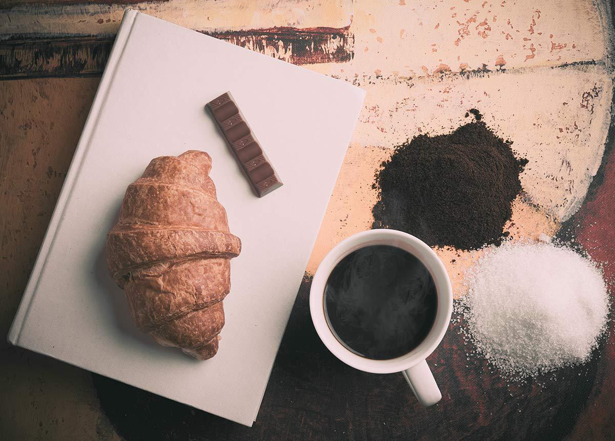 Croissant on a white cutting board with chocolate, coffee cup, cocoa powder, and sugar on a rustic table