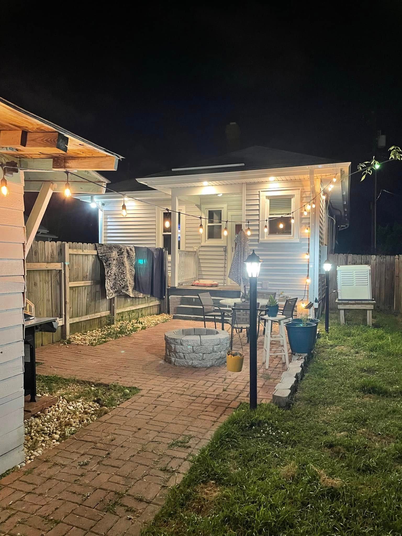 Small lit backyard shed at night, with brick path, grassy yard, and a fire pit in front.