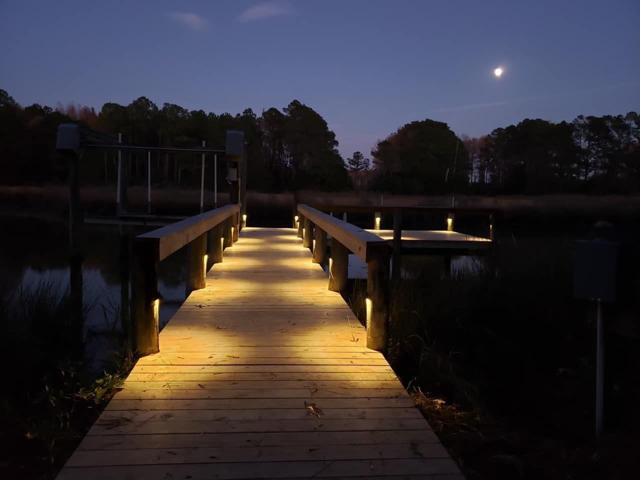 Illuminated wooden dock leading to a calm lake at dusk, with the moon above and trees in the background