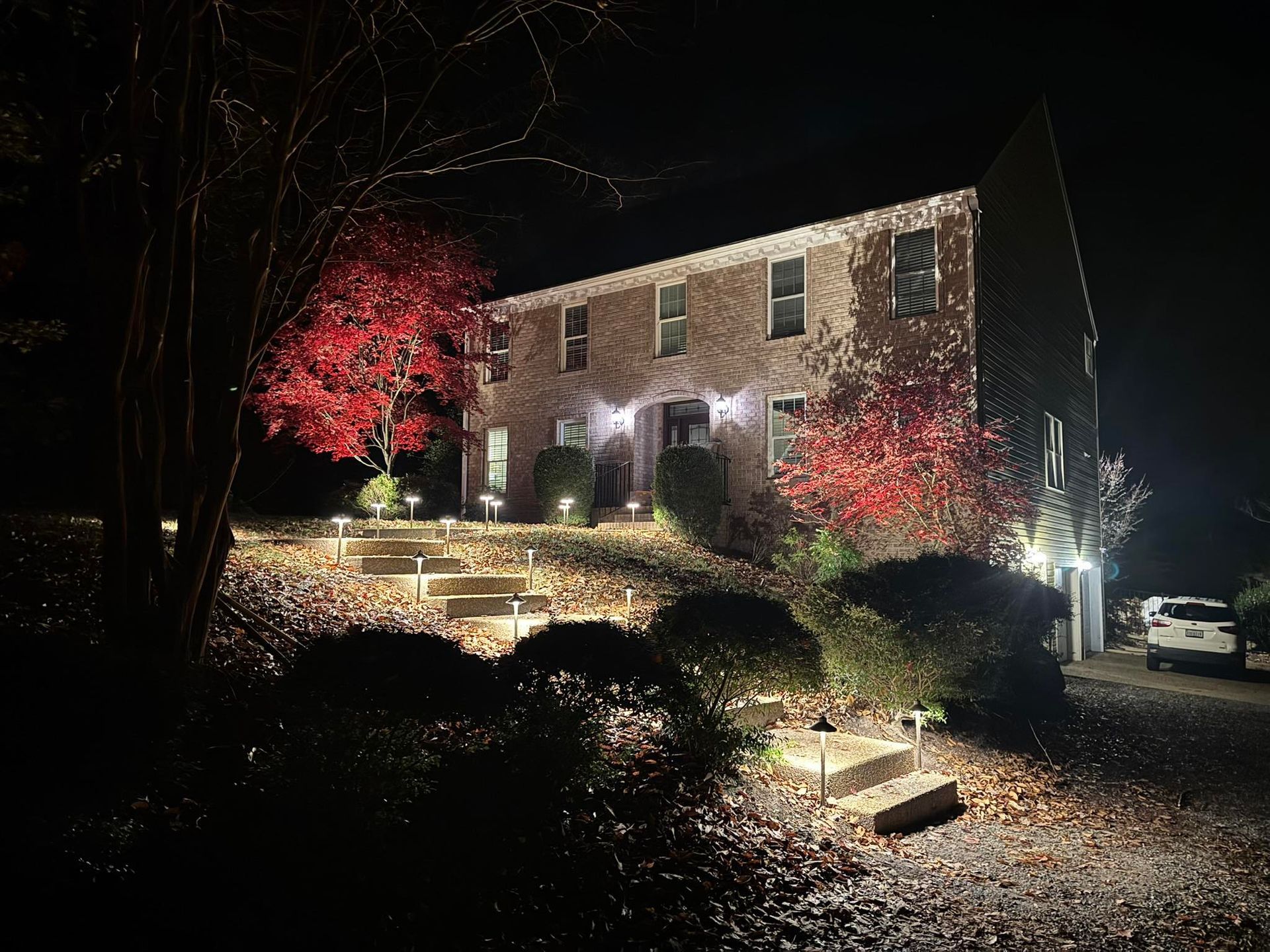 Nighttime view of a brick house lit by landscape lights, with red trees and stone steps in front.