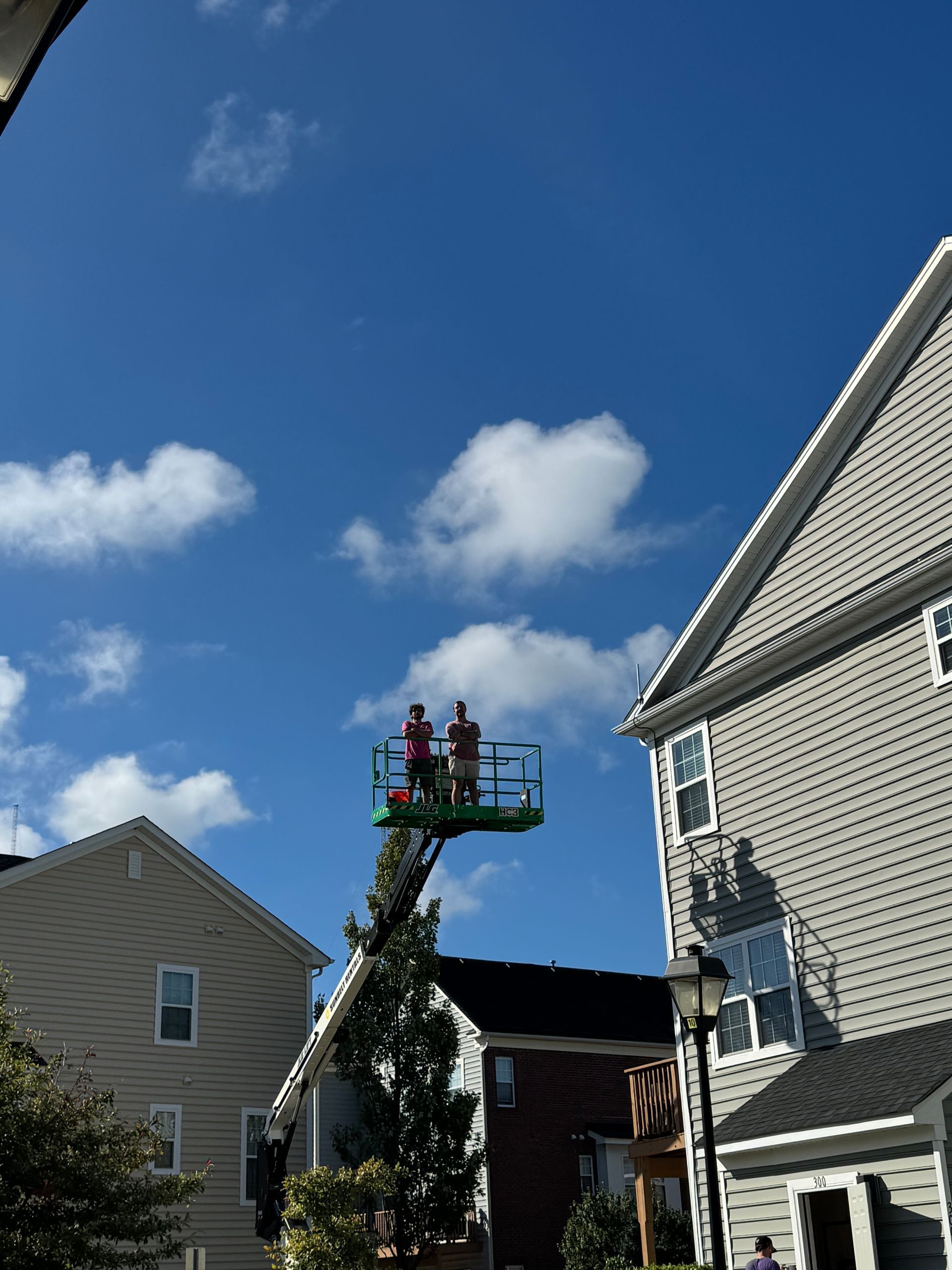 Workers in a green cherry picker trimming a tree between suburban houses under a blue sky