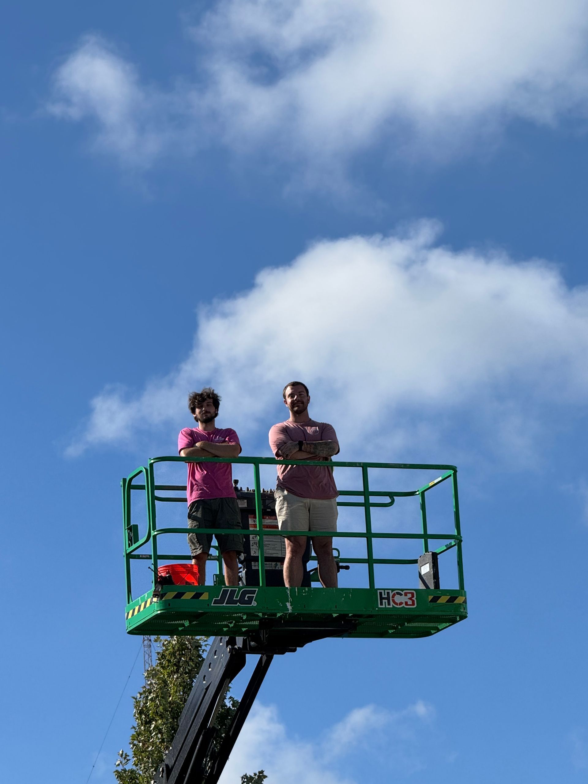 Two people standing on a green cherry picker against a blue sky with clouds