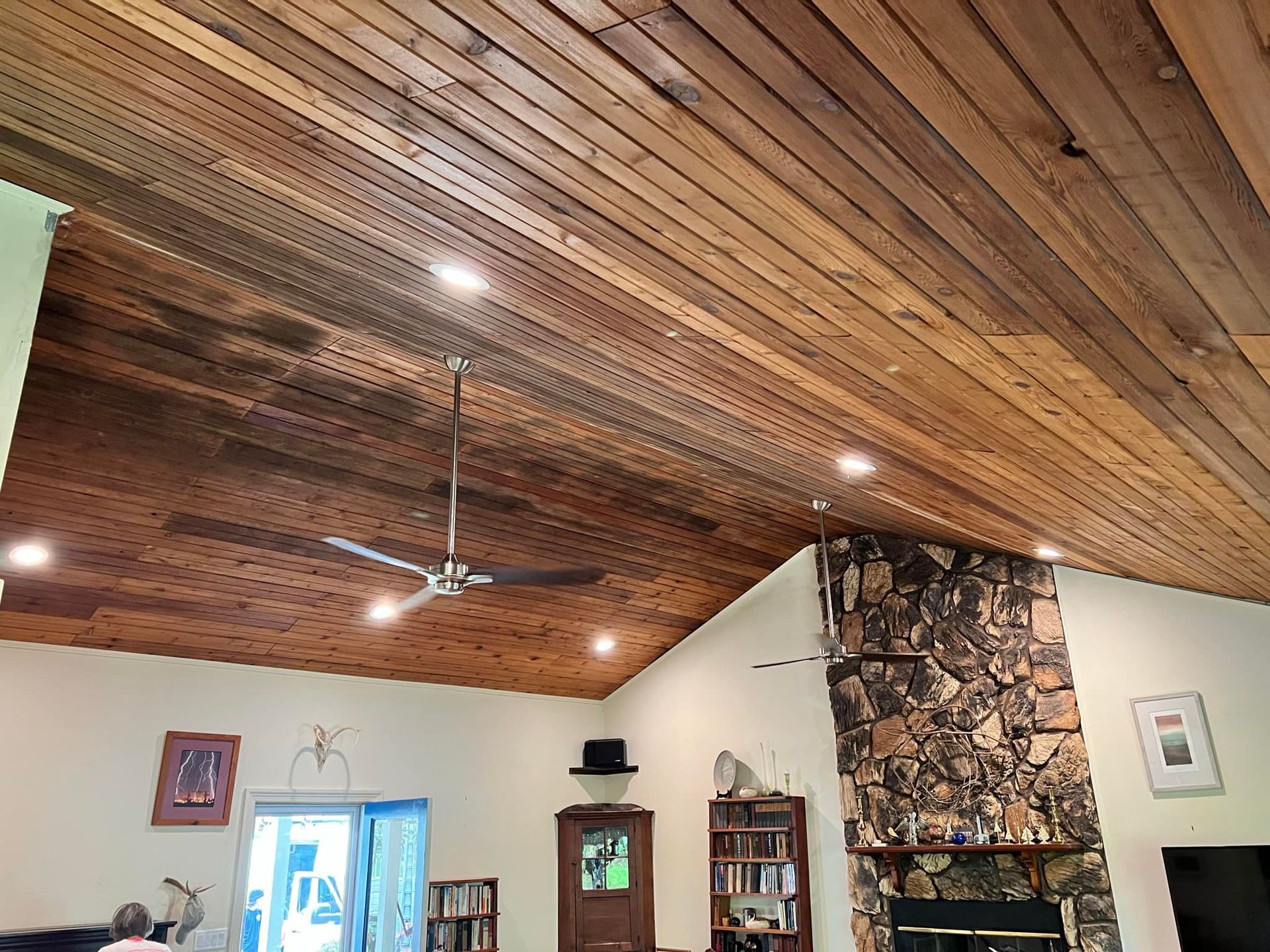 Wood-paneled vaulted ceiling with ceiling fan above a living room, stone fireplace, and bookshelves.