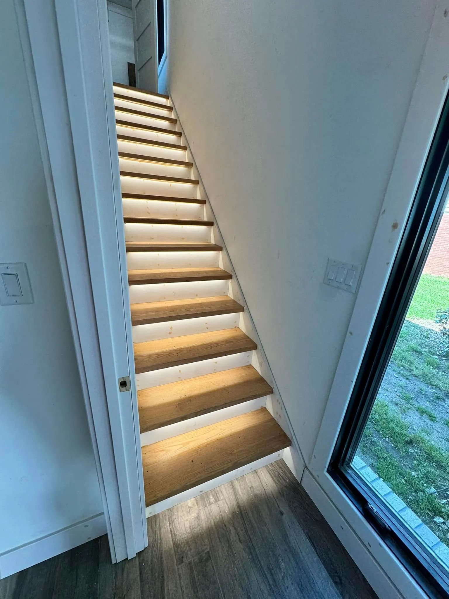 Narrow indoor staircase with illuminated wooden steps beside a tall window and white walls