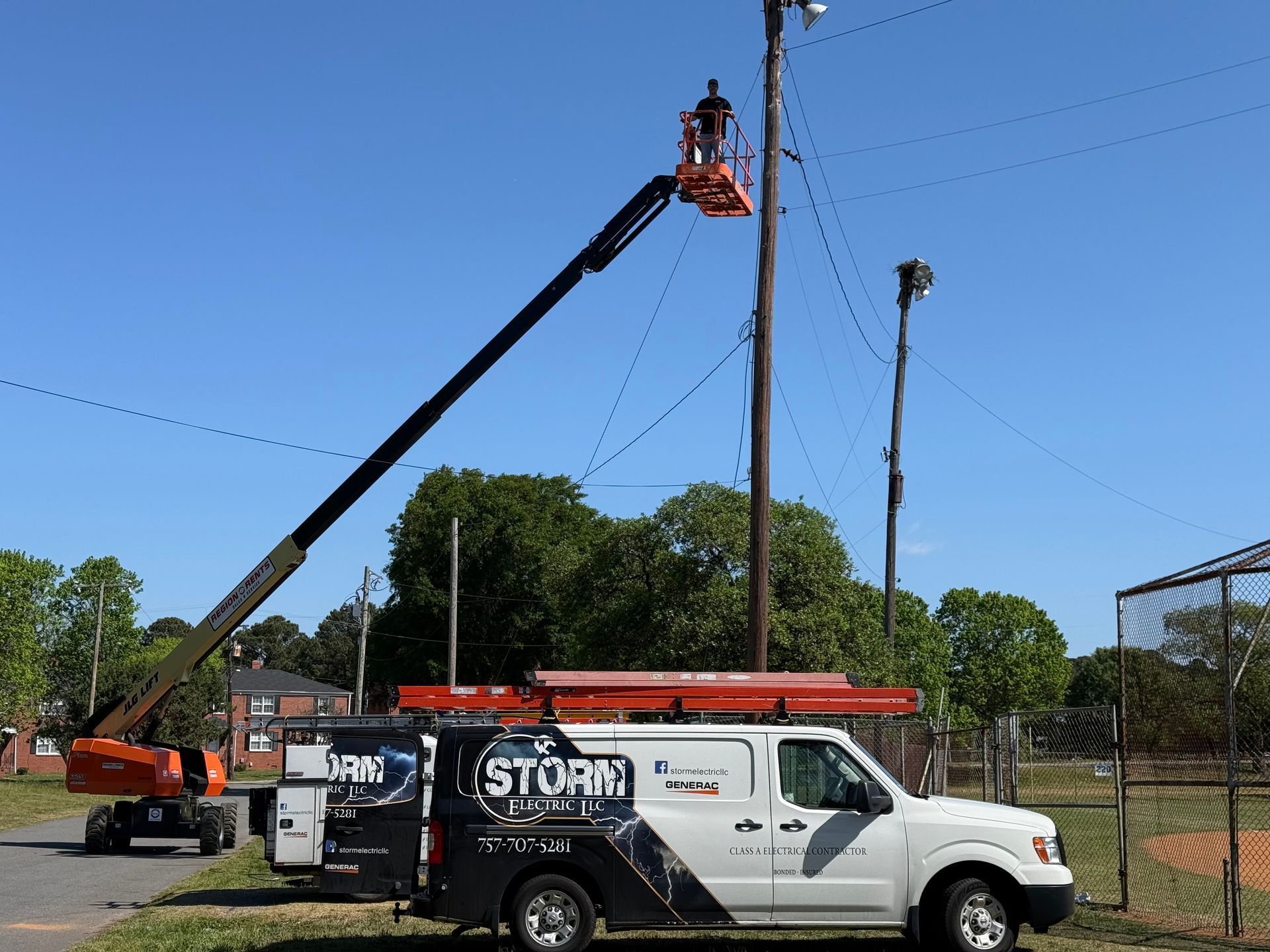 Utility worker in lift servicing a pole beside a black-and-white truck in a fenced field