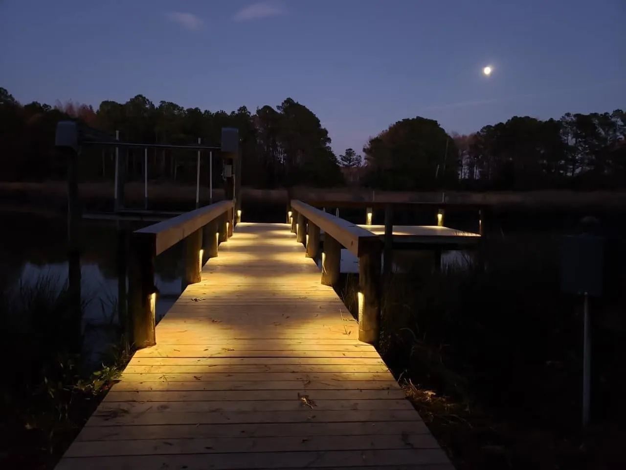 Illuminated wooden dock leading to a calm lake at dusk, with the moon above and trees in the background