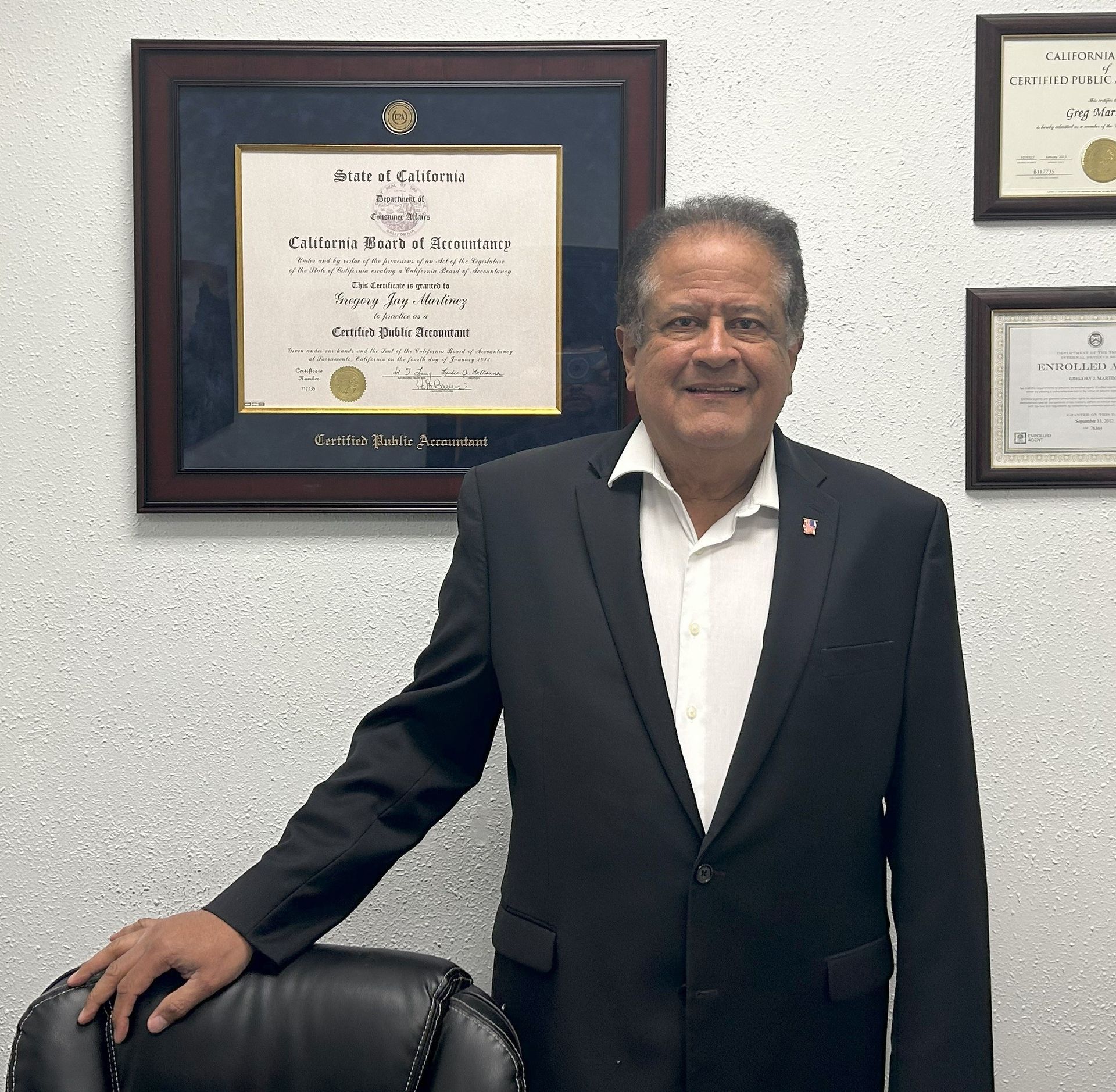 A man in a suit stands in front of a framed diploma