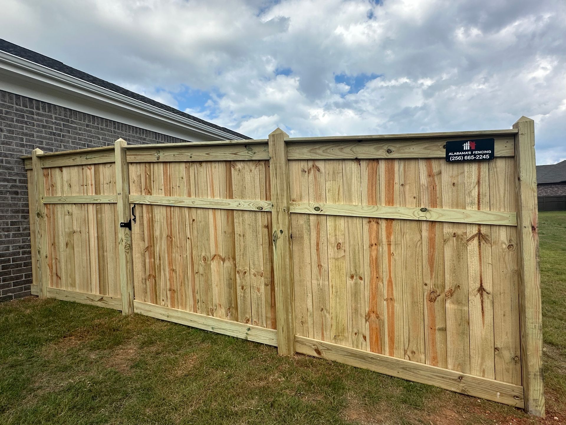 Wooden fence with gate next to a brick building and grass, under a cloudy sky.