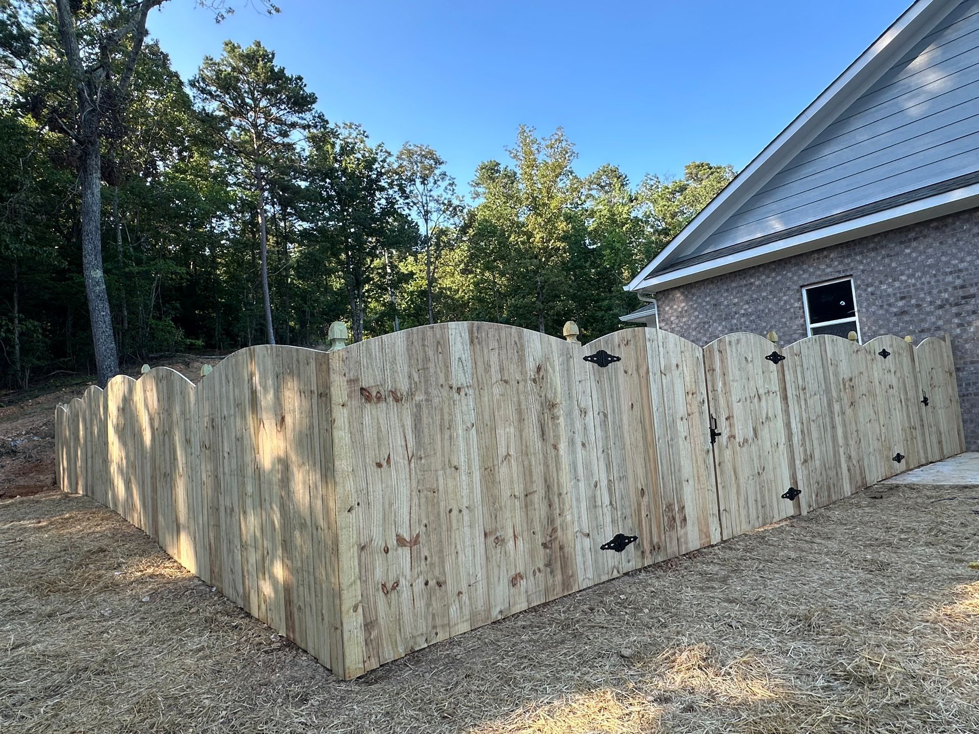 Wooden fence with arched tops bordering a brick building and a wooded area.