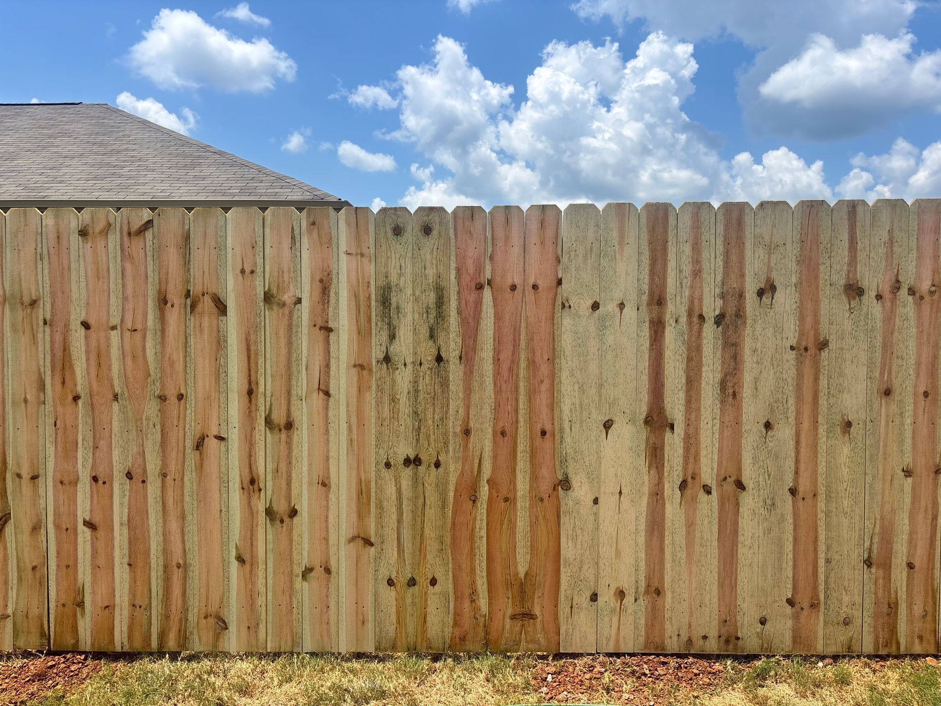 Wooden fence with a section of a house roof against a blue sky with clouds.