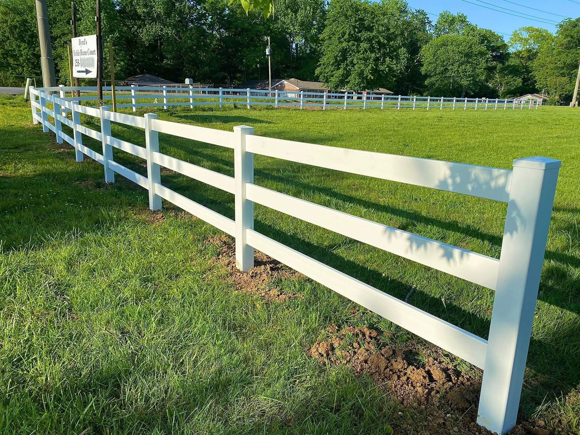 White three-rail fence in a grassy field. Blue sky and trees in background.