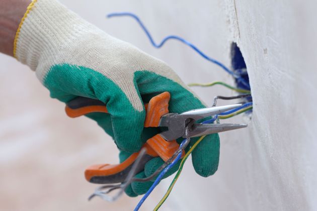 Hand with work glove using pliers to cut electrical wires in a wall.