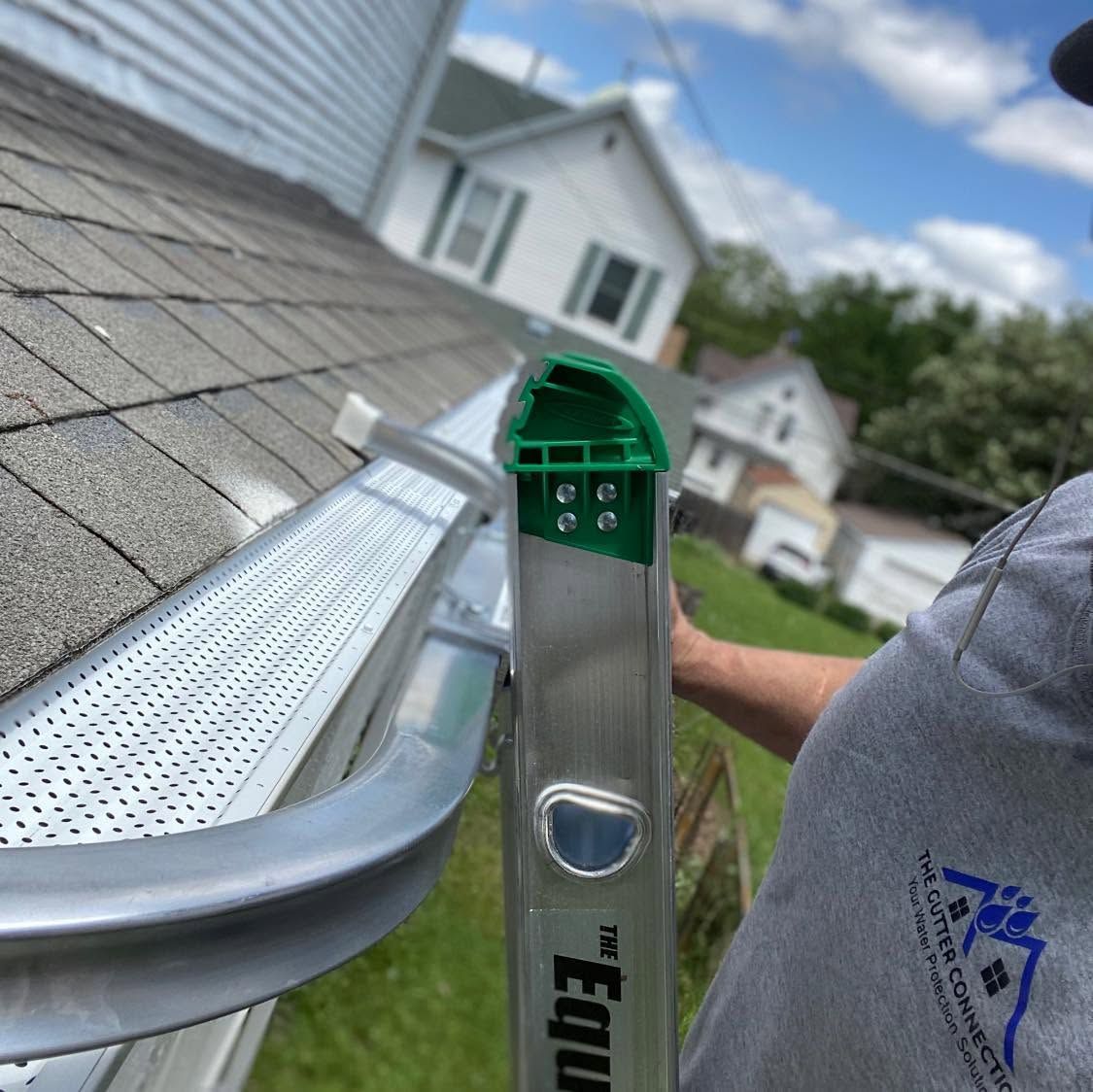 Gutter work on a roof, with a worker holding a ladder beside a white house and cloudy sky.