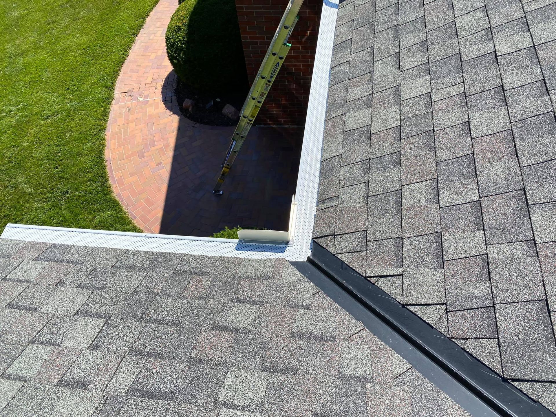 Roof shingles and gutter beside a grassy yard and a curved brick walkway, viewed from above