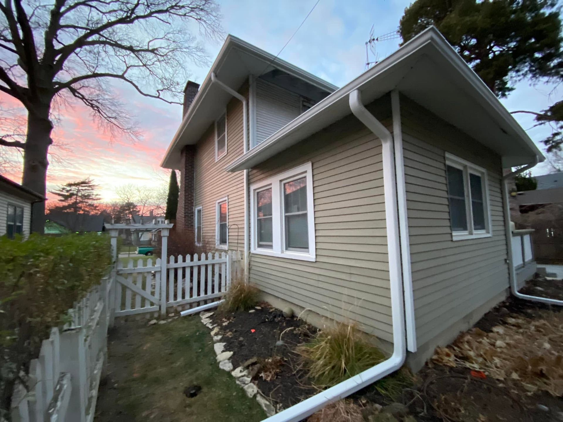 Side view of a tan house with white trim, a small porch, and a fenced yard at sunset.