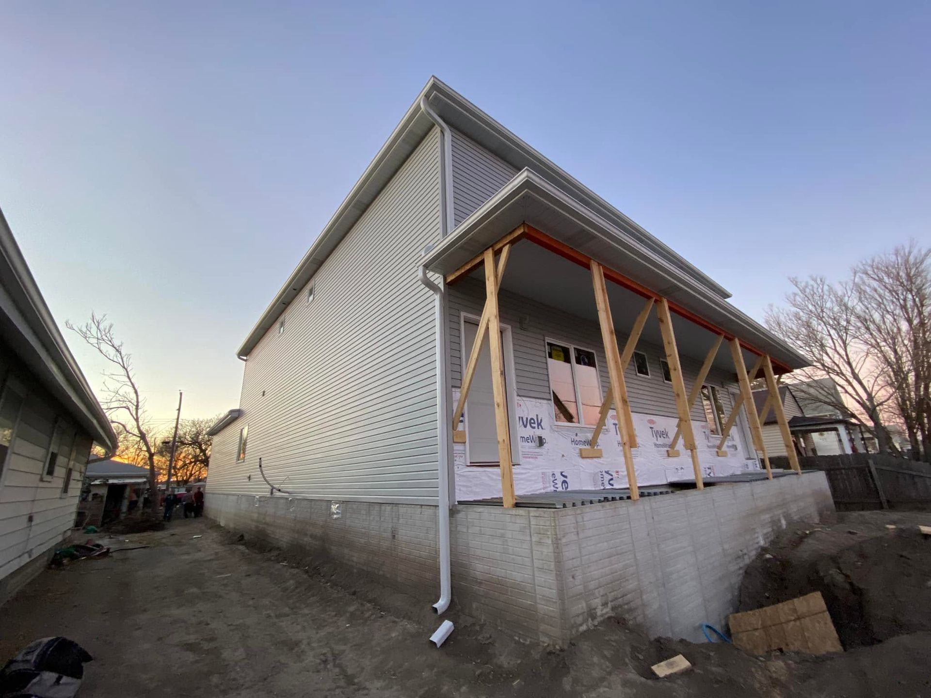 House under construction with exposed porch framing and white exterior wrap on a concrete foundation