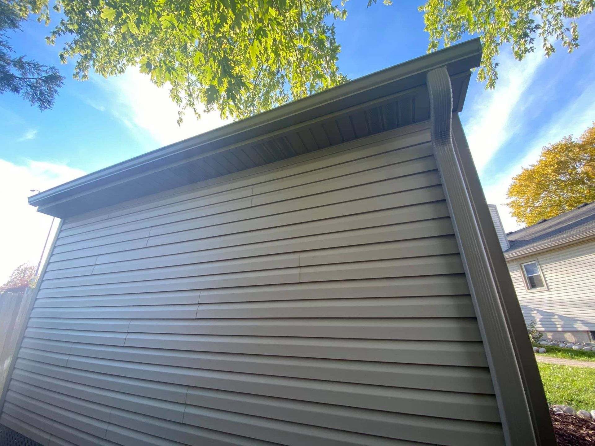 Low-angle view of a beige house exterior with dark trim beneath a bright blue sky and trees