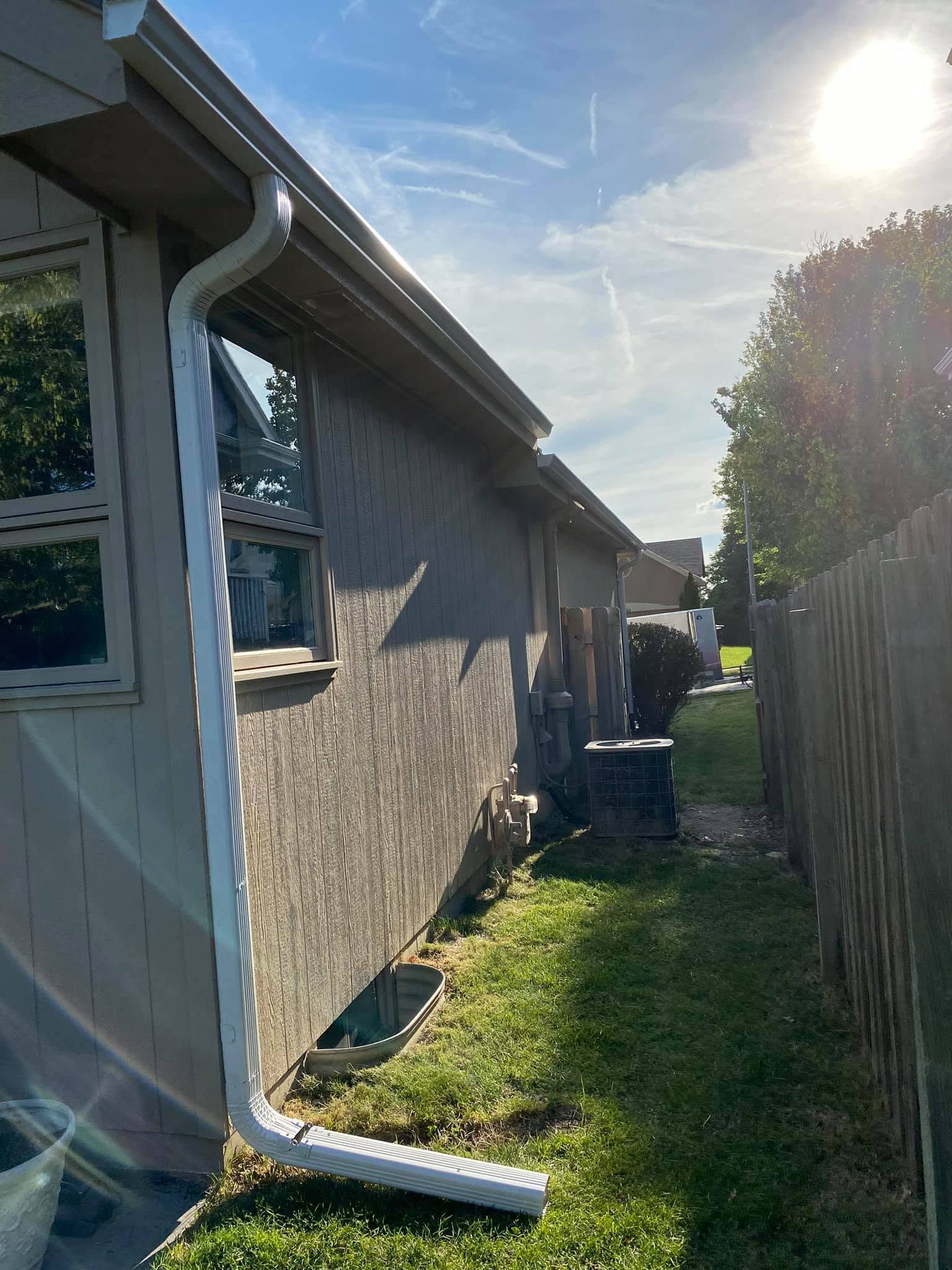 Side view of a house with a gutter, narrow grassy yard, and bright sun over a suburban fence