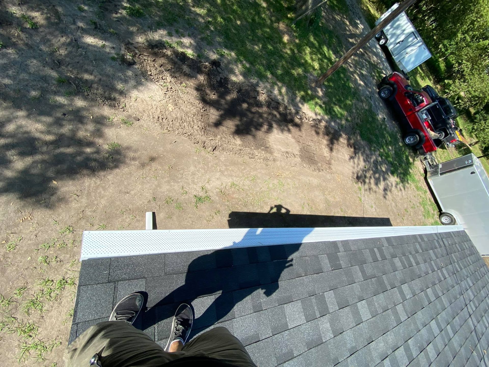 Person standing on a shingled roof, looking down at a driveway with a red car and trees nearby.