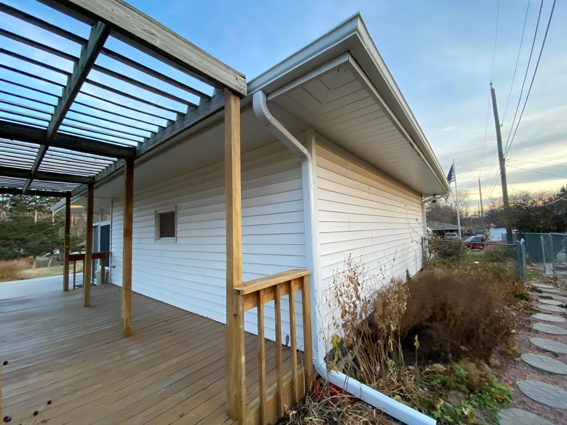 Wooden deck beside white house with pergola and shrubs at sunset