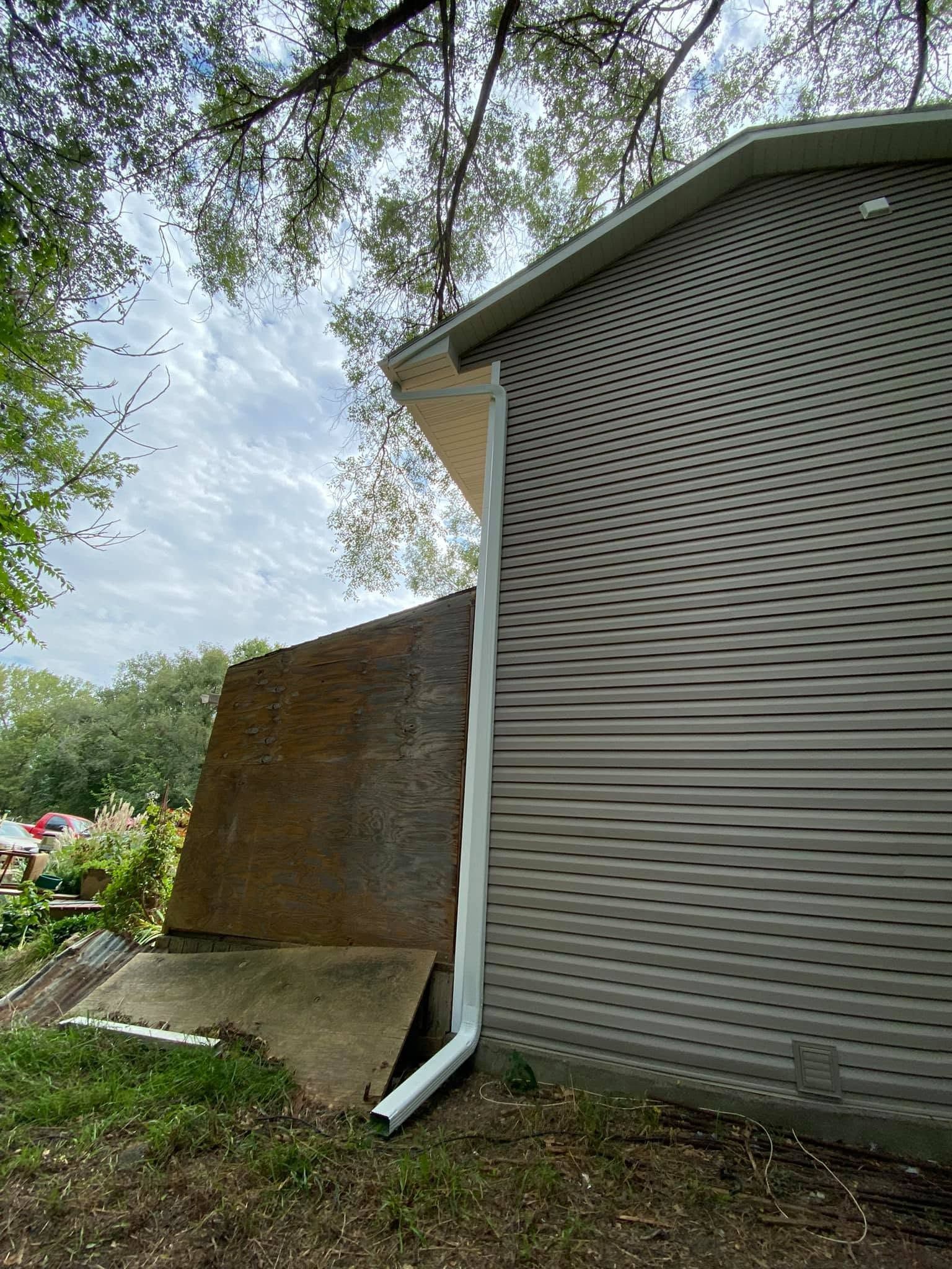 Side of a house with gray siding, a leaning rusted panel, and a white downspout under trees.