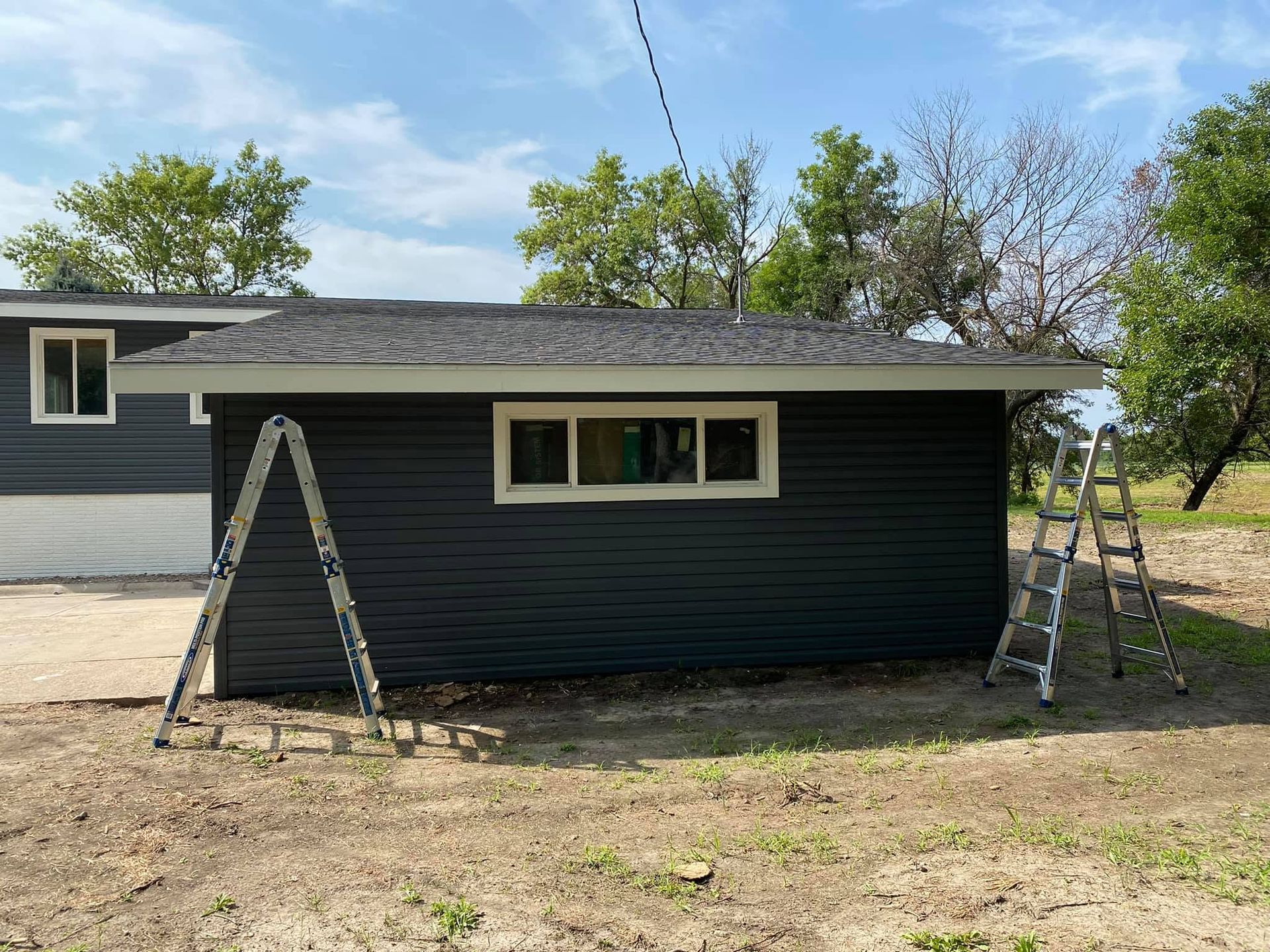 One-story dark-sided house under renovation, with two ladders leaning against the exterior.