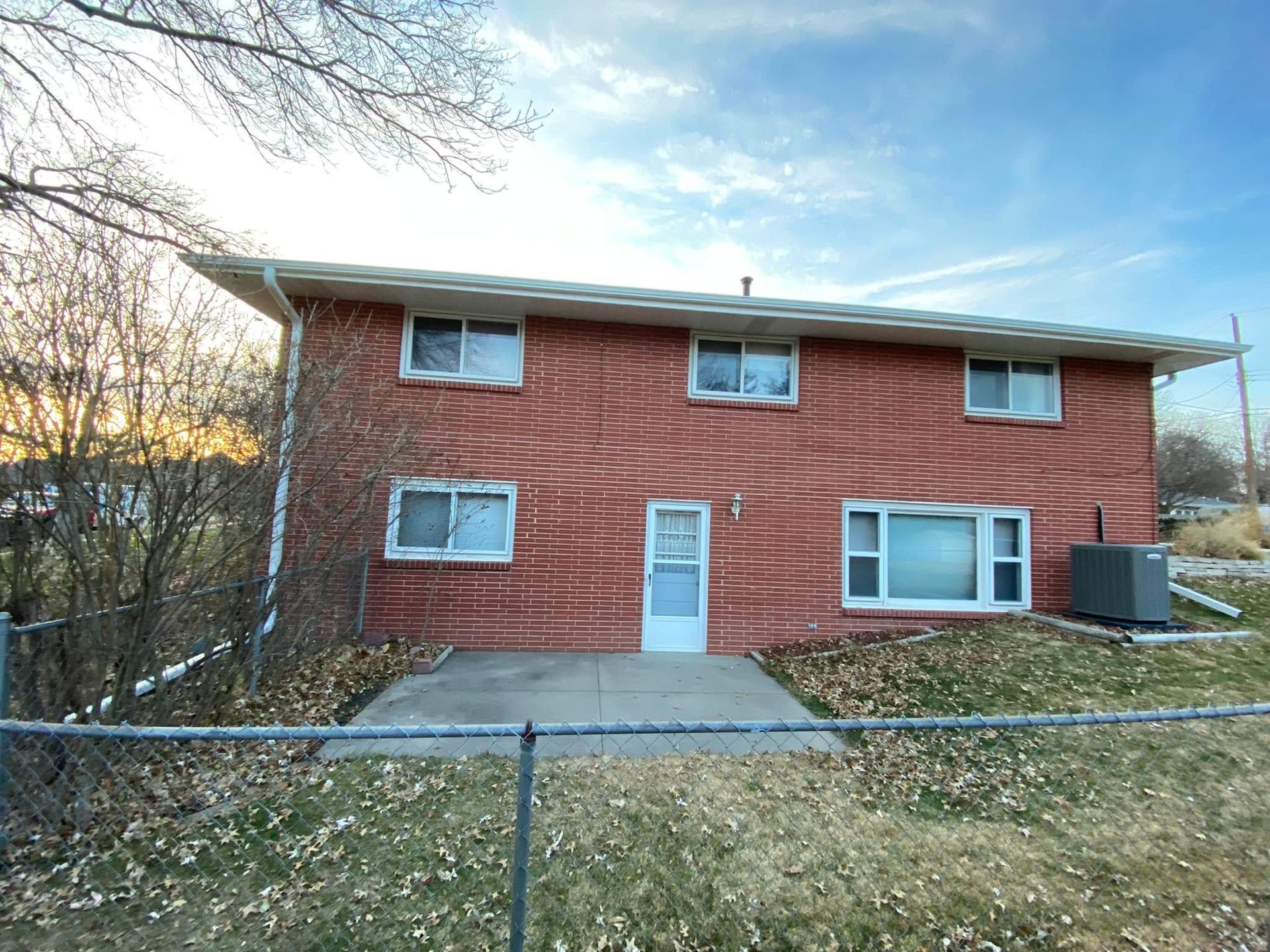 Red brick two-story house with white trim, front door, driveway, and chain-link fence in front.