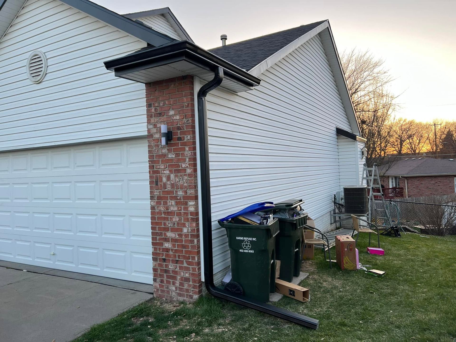 Side of a white house with brick column, garage, trash bins, and lawn at sunset