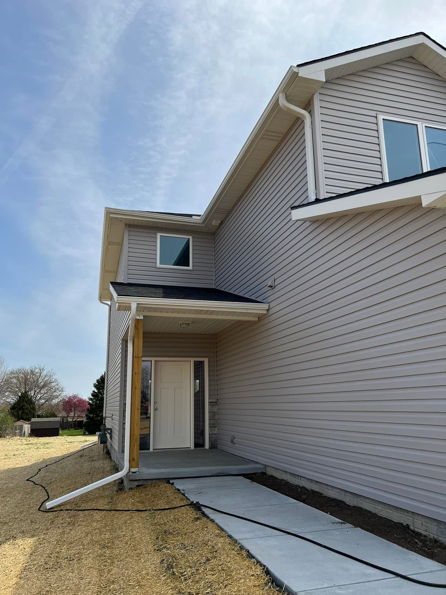 Side view of a beige house exterior with white trim, a small porch, and a concrete walkway on a grassy lot.