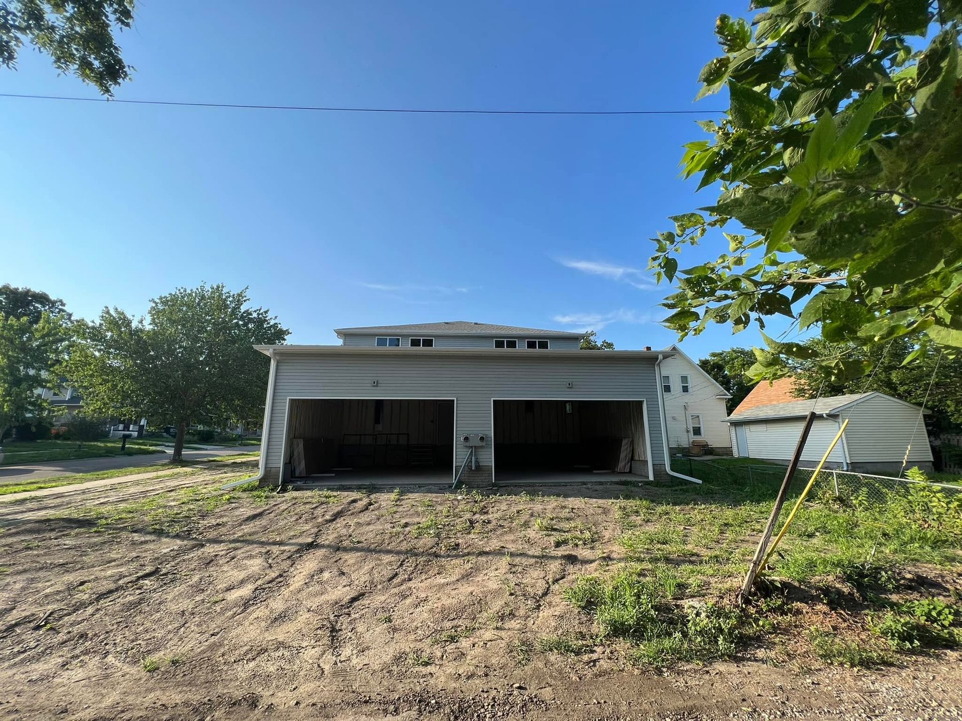 Two-car garage with open bays on a dirt lot, under a clear blue sky, with trees and a small shed nearby