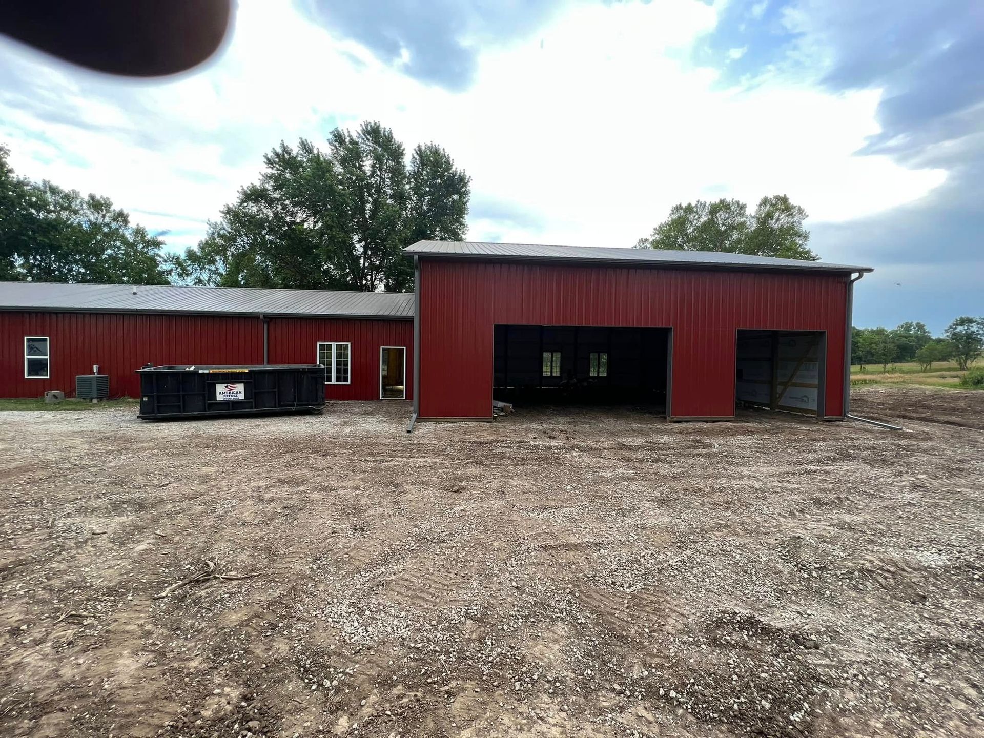 Red metal barn with open garage bays beside a gravel lot under a cloudy sky