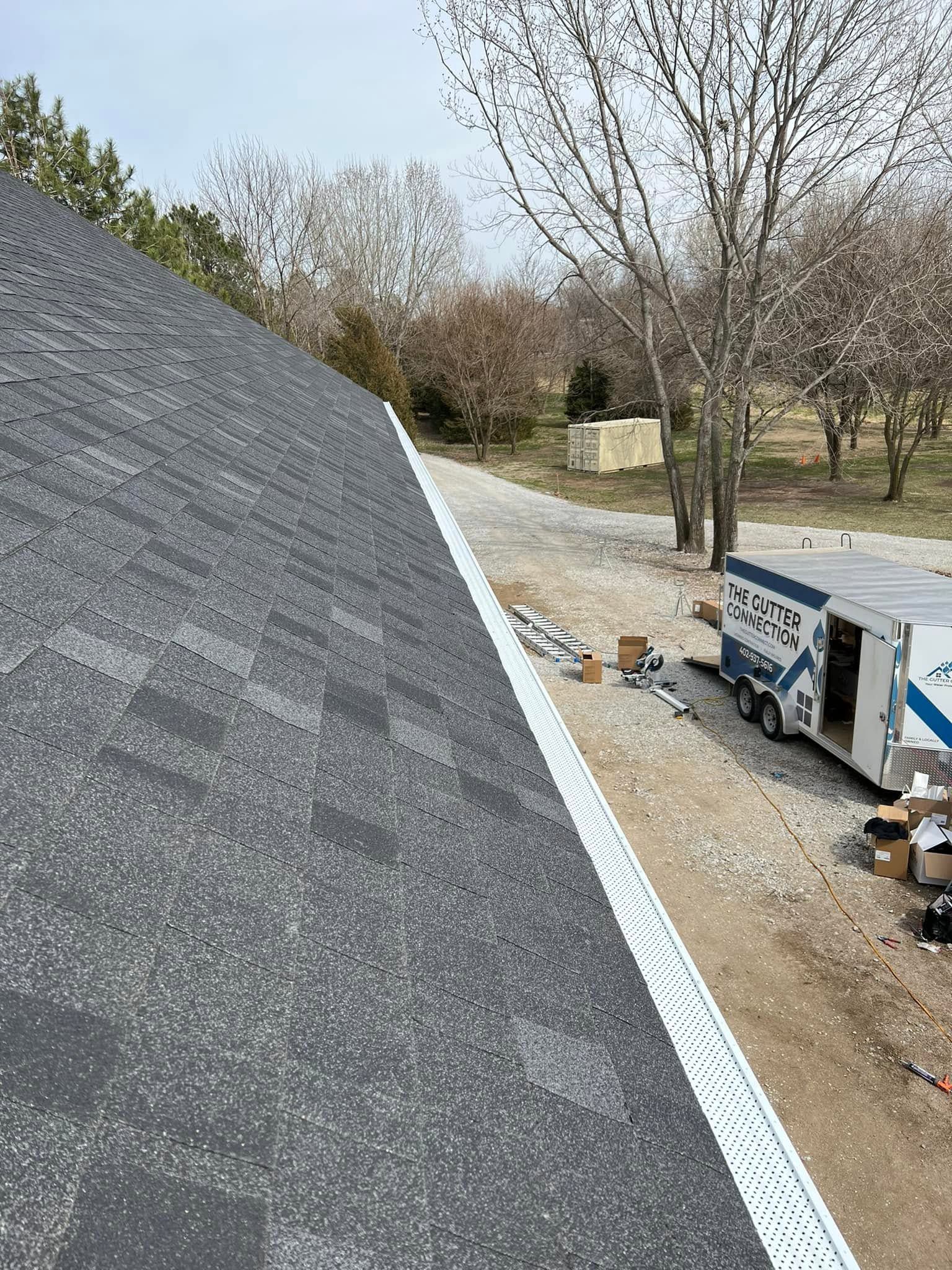 Roof shingles in the foreground with trees and a parked utility truck below on a gravel driveway.
