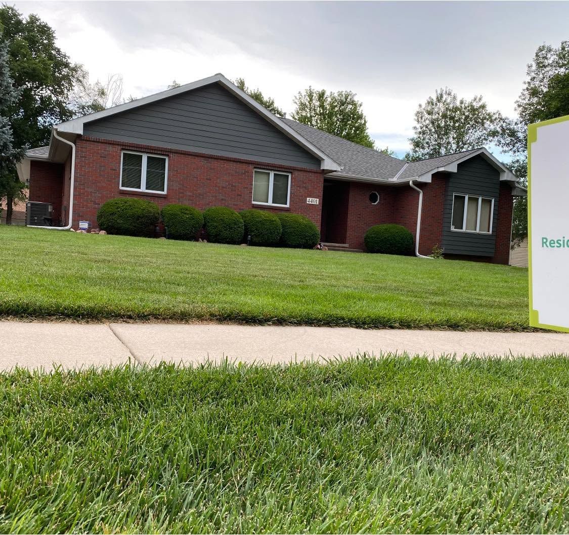 Brick ranch house with gray roof and trimmed hedges across a green lawn on a suburban street