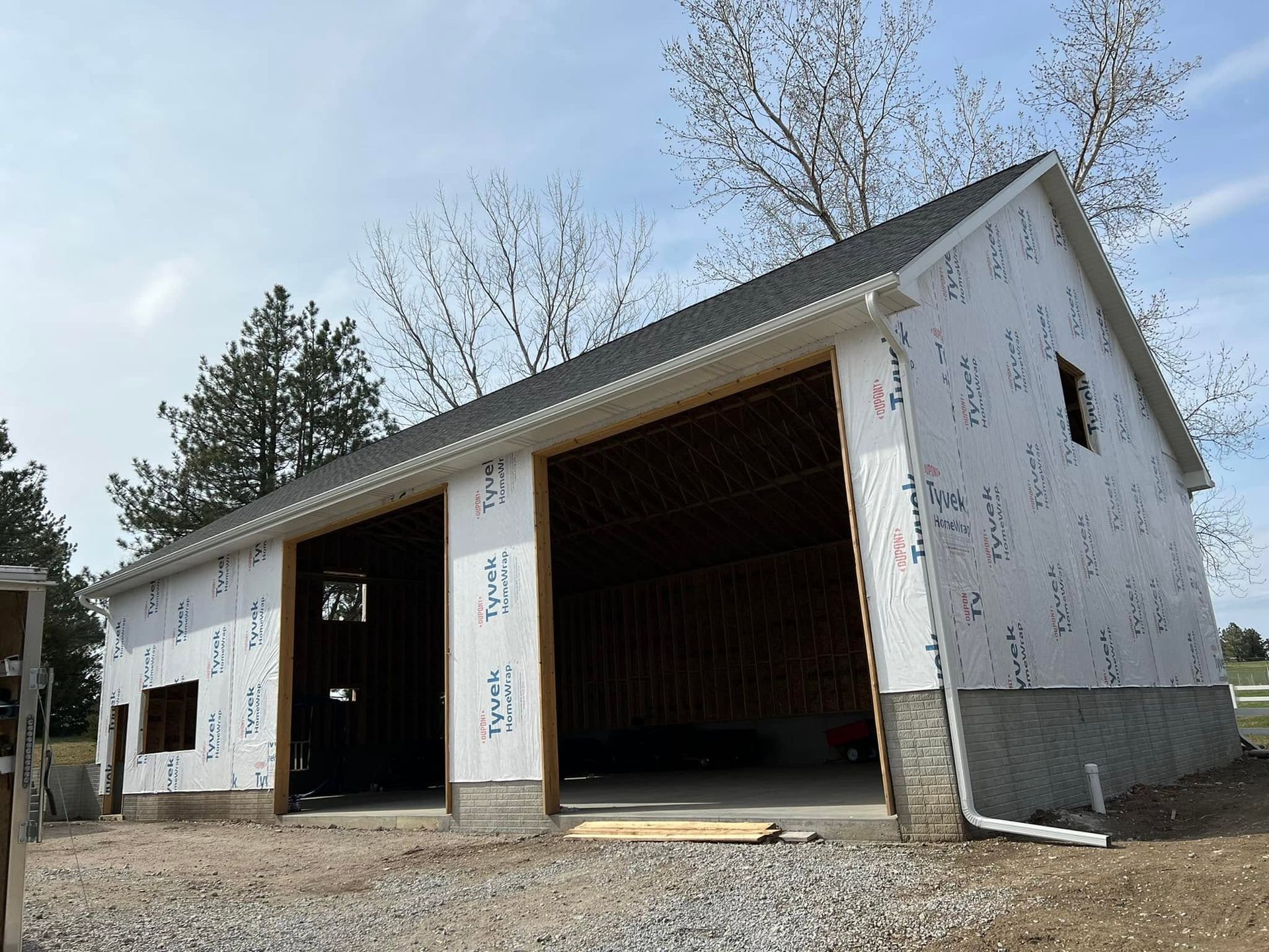Unfinished white garage or shed with open bays and a dark roof on a gravel lot under a cloudy sky